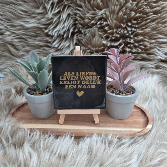 Keramisch zwart tegeltje met gouden tekst “Als liefde leven wordt krijgt geluk een naam” op houten standaard met twee plantjes – decoratie of cadeau.