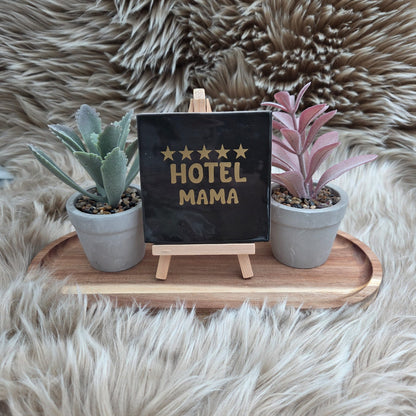 Zwart keramisch tegeltje met gouden tekst en vijf sterren “Hotel Mama” op houten standaard, met twee kleine planten – grappig moederdagcadeau.