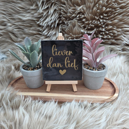 Keramisch tegeltje zwart met gouden tekst “Liever dan lief” en hartje op houten standaard tussen twee plantjes – romantische woondecoratie.